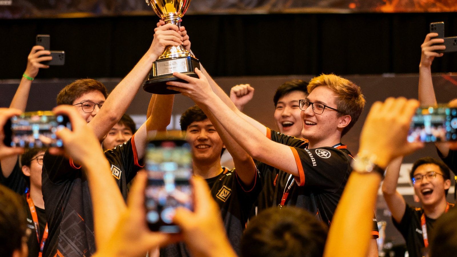DOTA2社区锦标赛颁奖环节，获胜队伍高举奖杯，周围观众纷纷举起手机拍照记录这一荣耀时刻